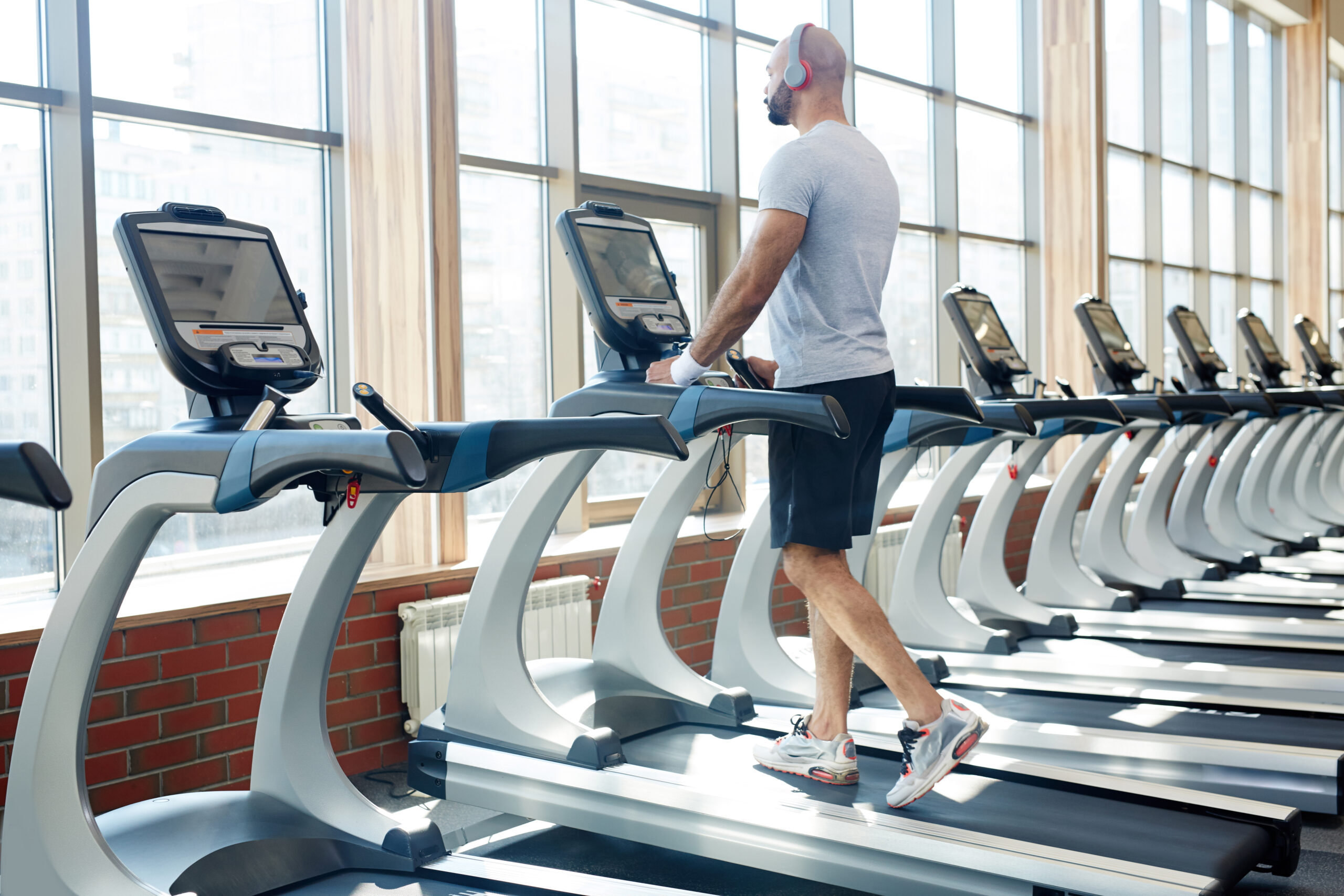 Back view portrait of muscular sportsman running on treadmill listening to music alone in empty sunlit gym